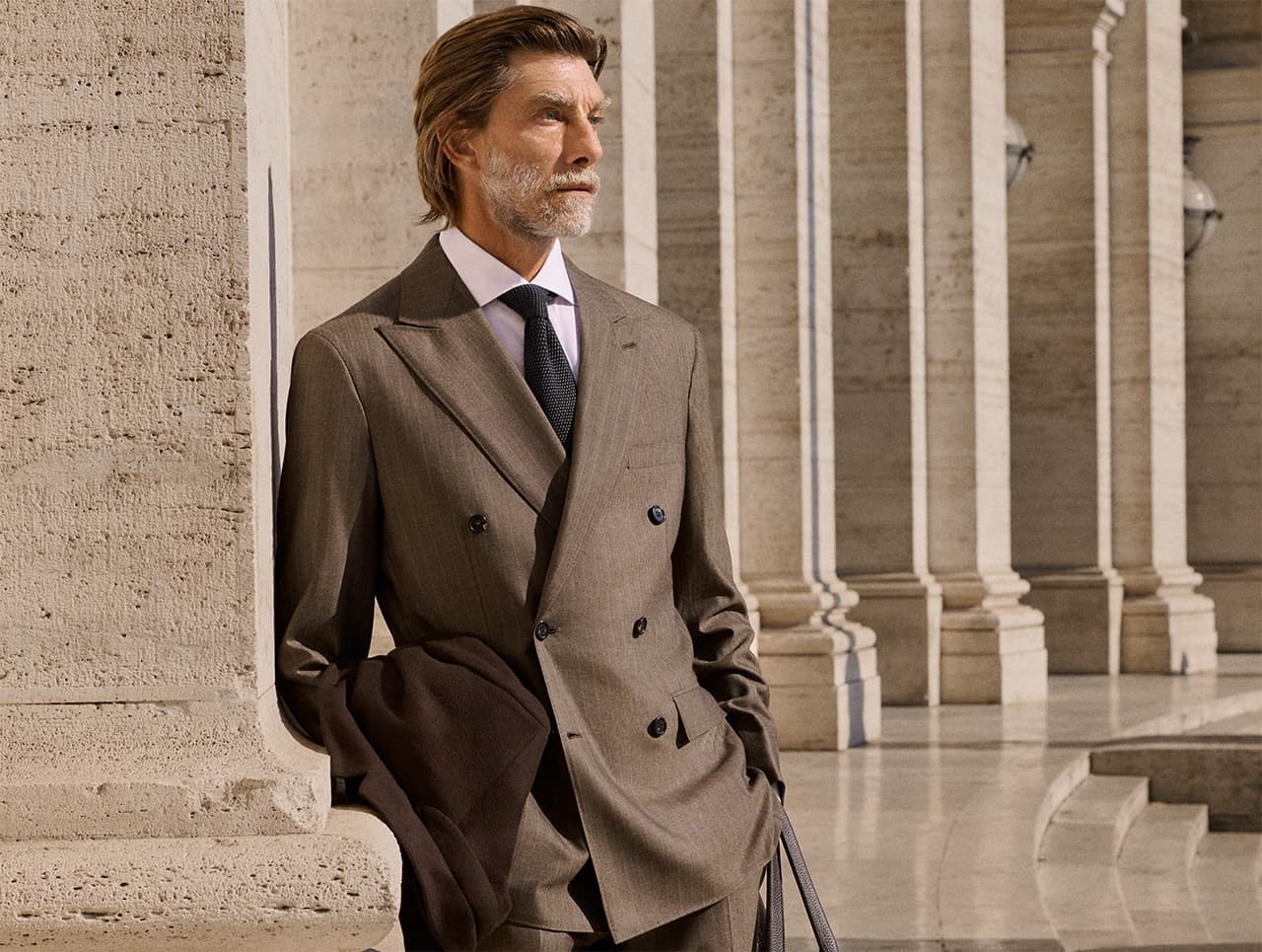 Distinguished man with gray beard in tan Brioni double-breasted suit and navy tie standing by stone columns in classical architecture.