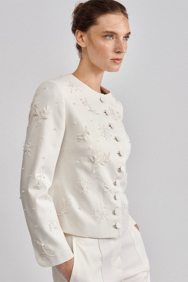 Woman in profile wearing Brioni white embellished jacket with snowflake and floral beading against light gray background.