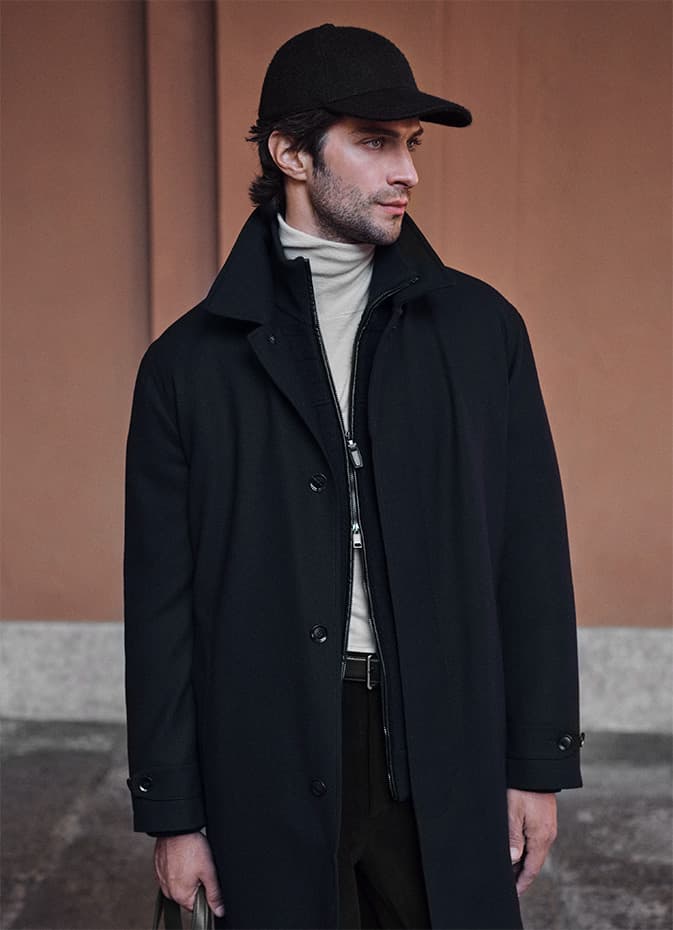Person in Brioni black coat and cap with white turtleneck against terracotta background.