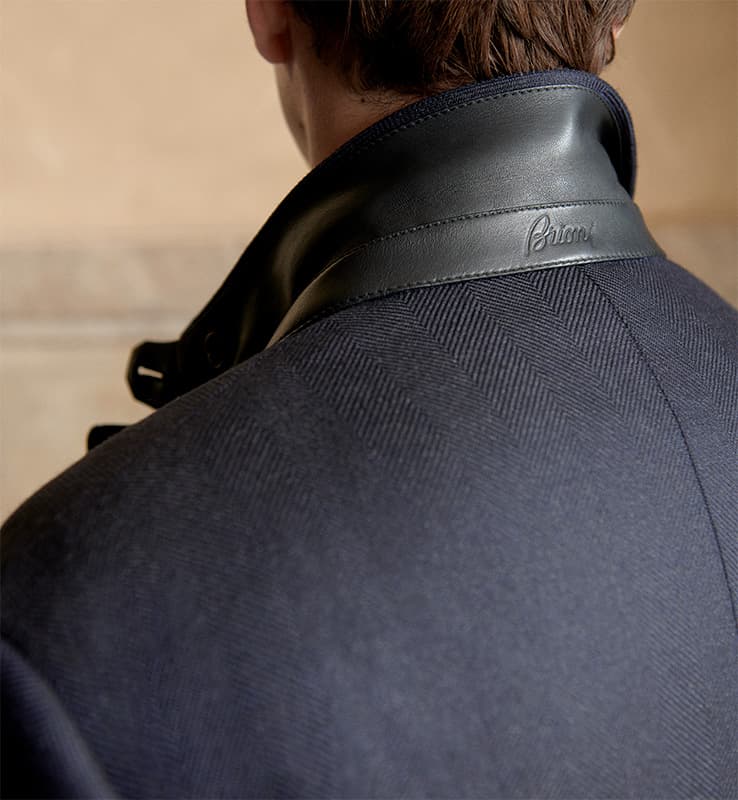Close-up of a Brioni navy blue coat with leather collar detail featuring an embossed Brioni logo against a neutral background.