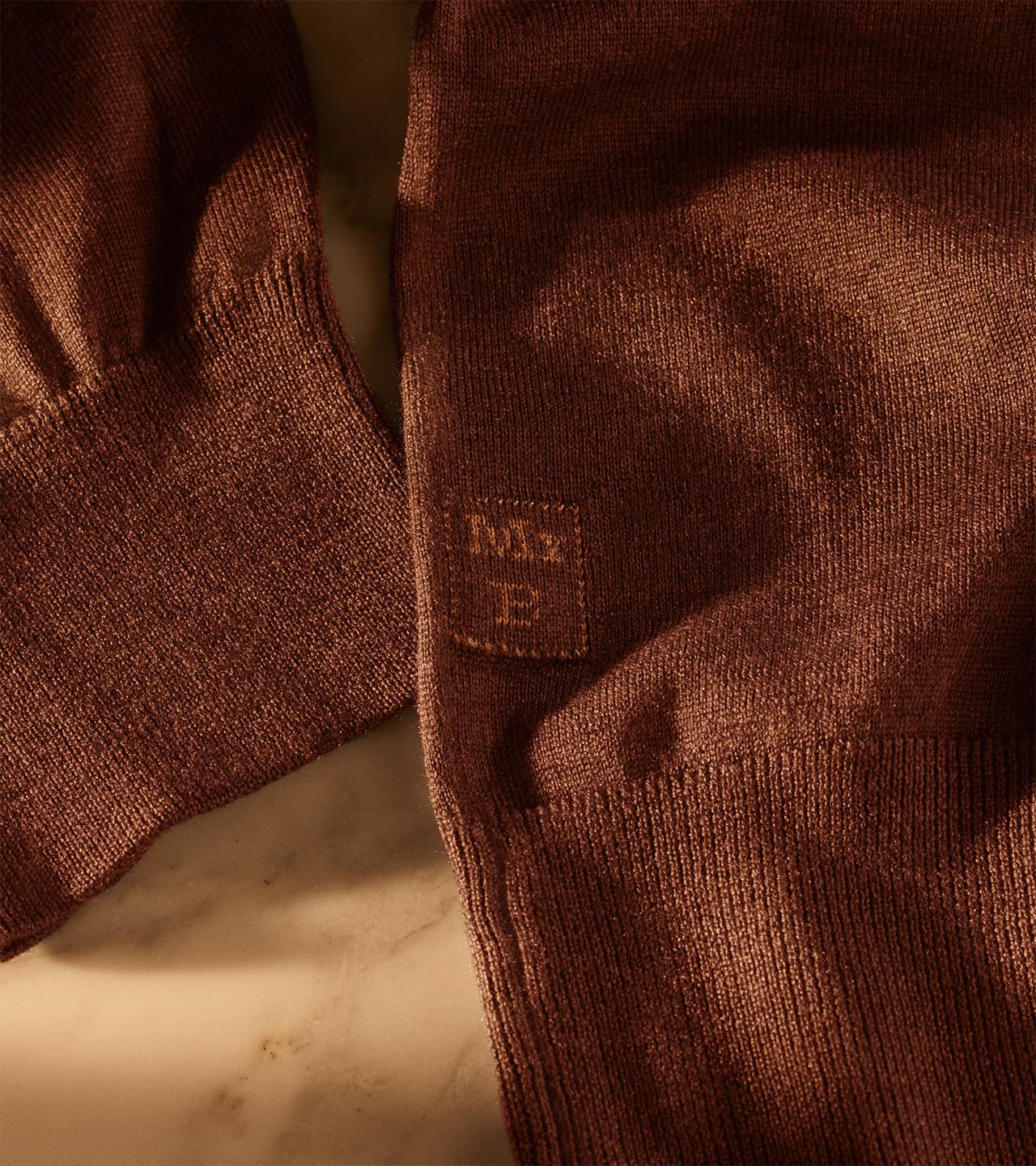 Close-up of Brioni rust-colored knit sweater showing textured fabric detail and ribbed cuffs with subtle embroidered logo.
