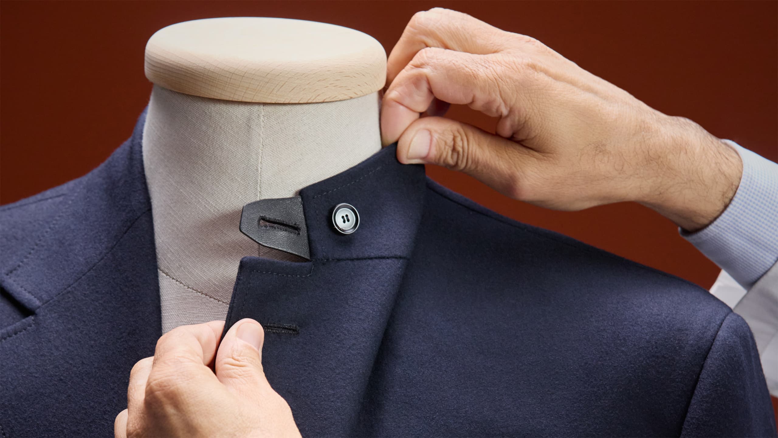 Hands adjusting the collar of a Brioni navy blue coat on a mannequin, showing tailored craftsmanship against a red background.