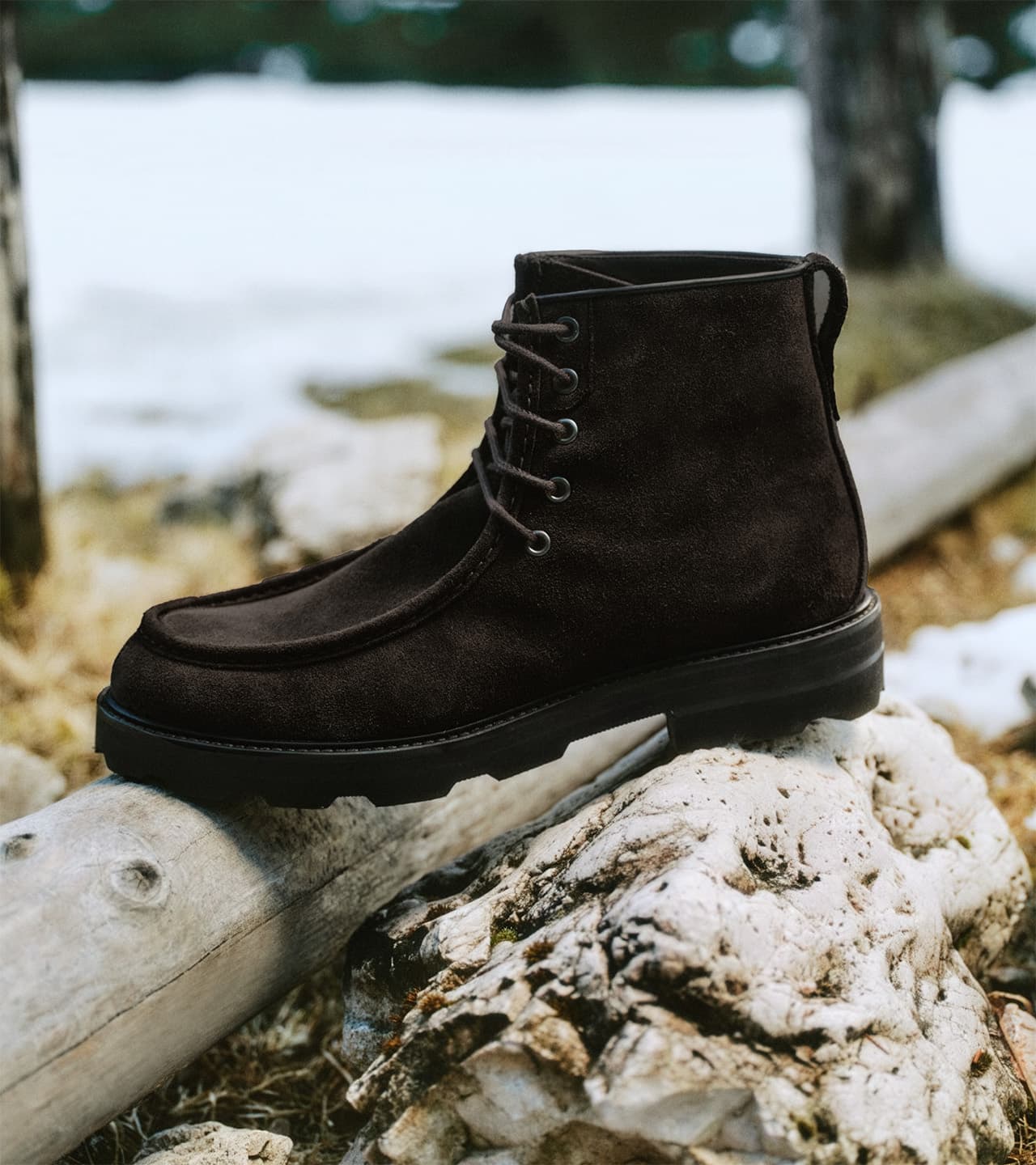 Brioni dark brown suede ankle boot with laces displayed on a weathered log in a snowy outdoor setting.