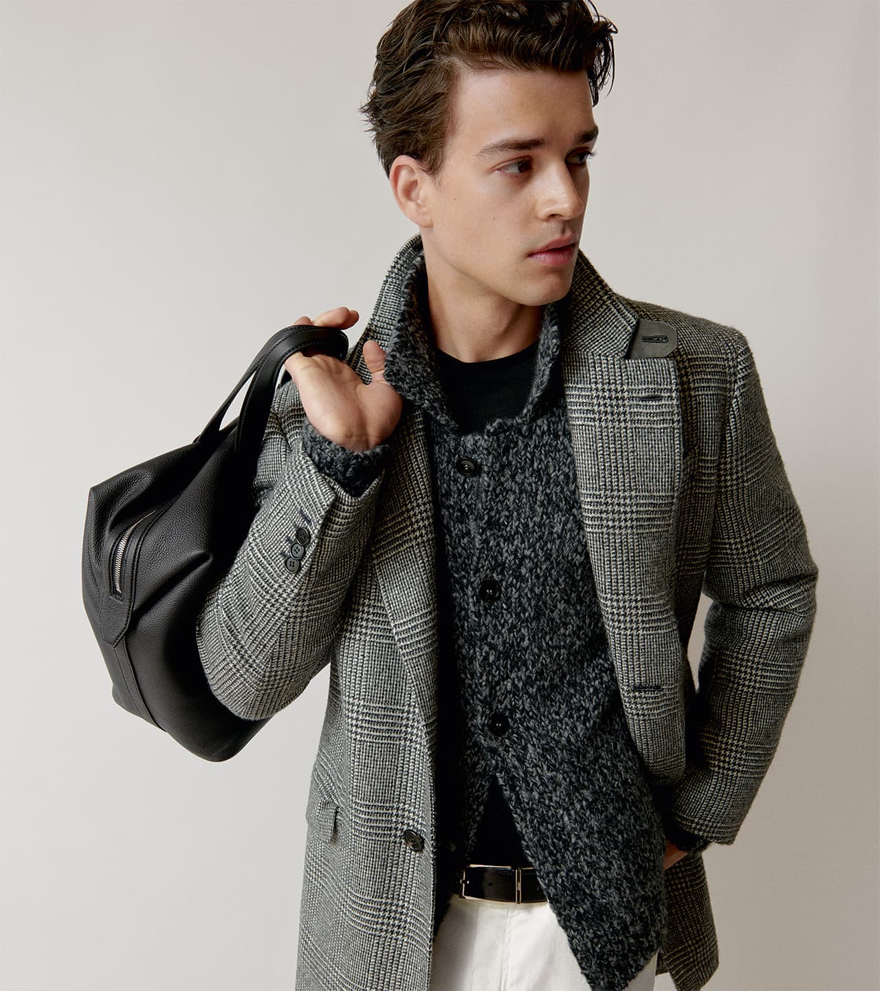 Young man in Brioni plaid blazer and textured coat carrying a black leather bag against neutral background.
