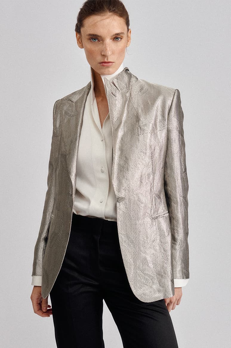 Woman wearing Brioni silver textured blazer over white blouse and black pants against gray background in professional portrait.