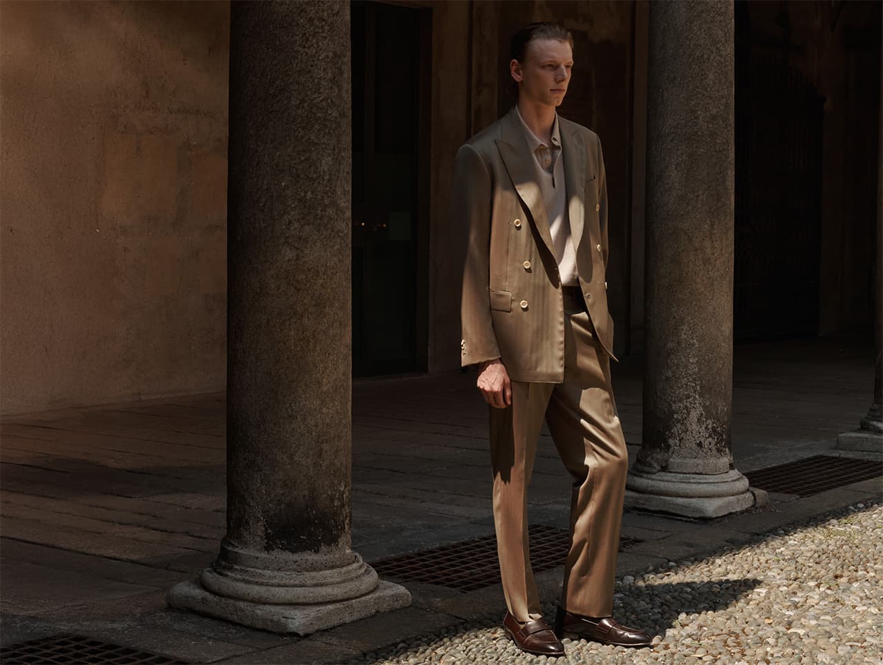 Man in Brioni beige double-breasted suit standing between stone columns in architectural setting with dramatic lighting.
