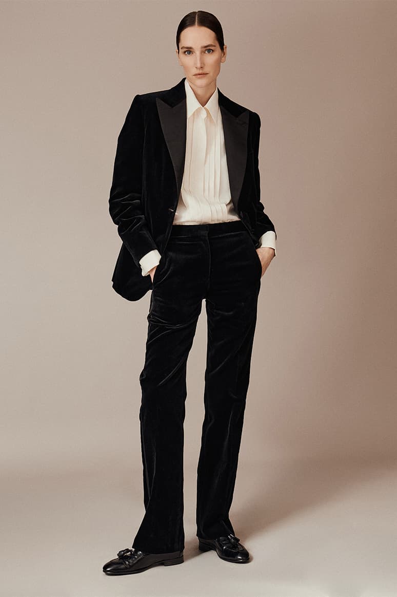Woman wearing Brioni black velvet tuxedo suit with white shirt and black shoes against beige background in formal portrait pose.