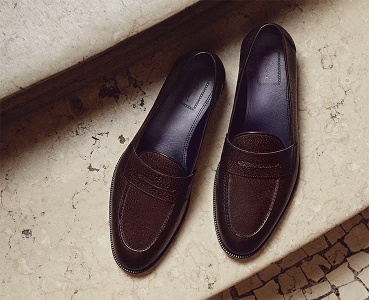 Pair of elegant Brioni dark brown leather penny loafers on a cream-colored stone surface with subtle texture