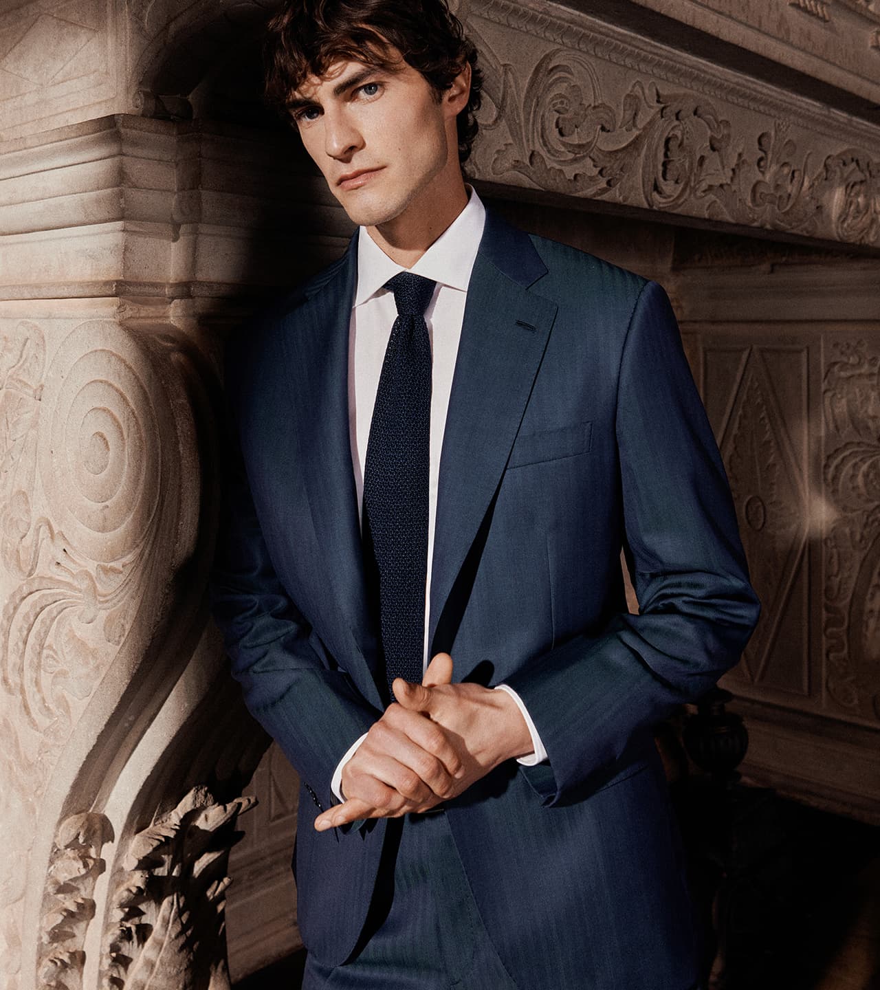 Man in navy blue Brioni suit with white shirt and dark tie standing against ornate stone architecture with carved details.
