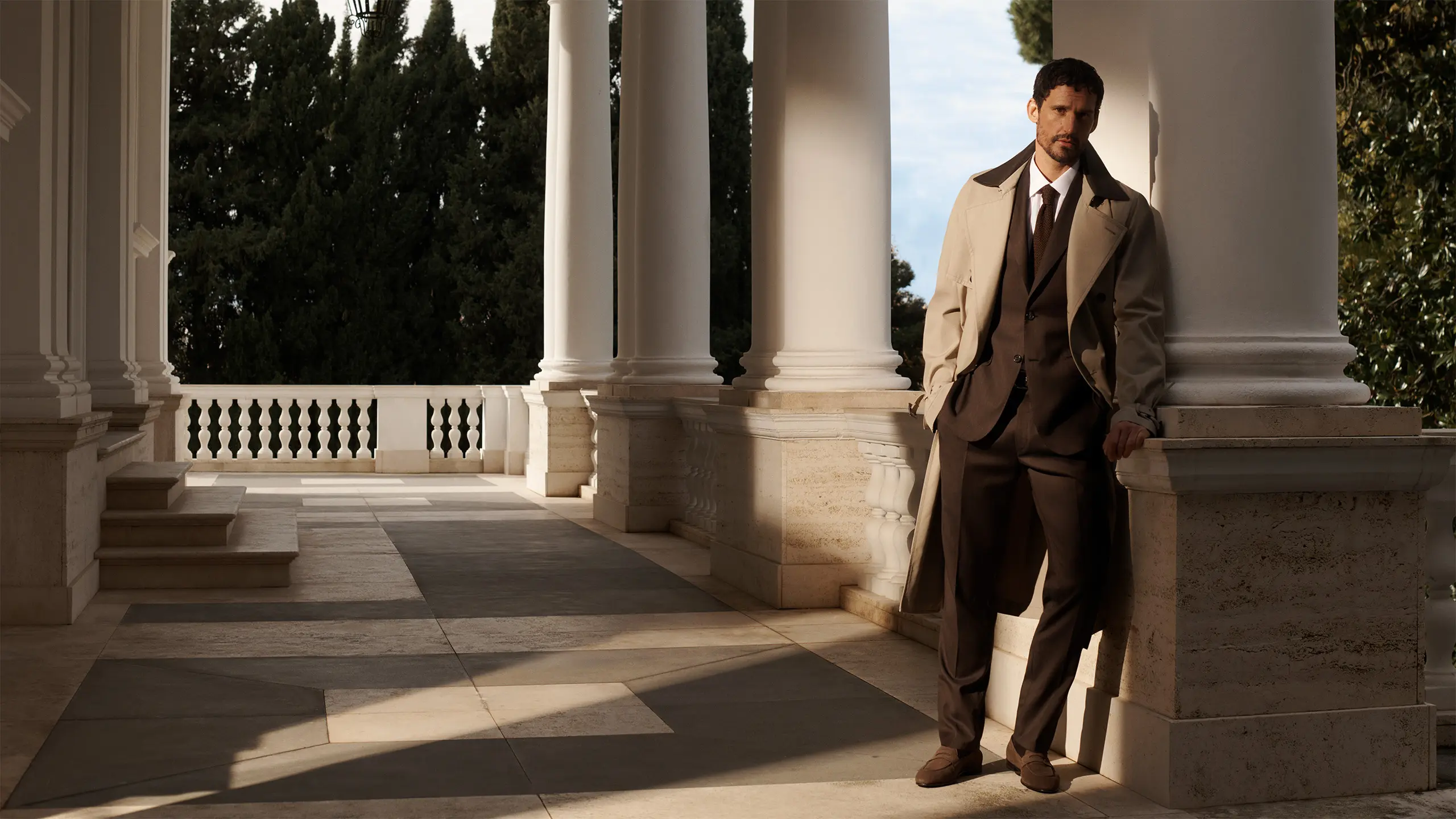 Man in a Brioni beige suit and tie leaning against white column on elegant terrace with balustrade and trees in background.