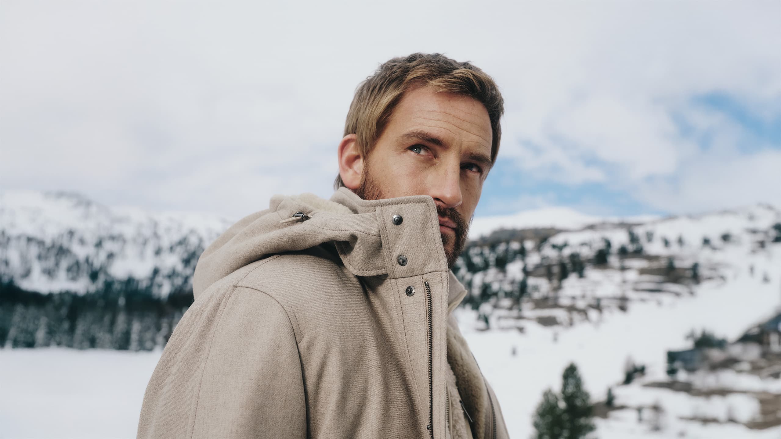 Person in Brioni beige winter coat against snowy mountain landscape with cloudy sky background.