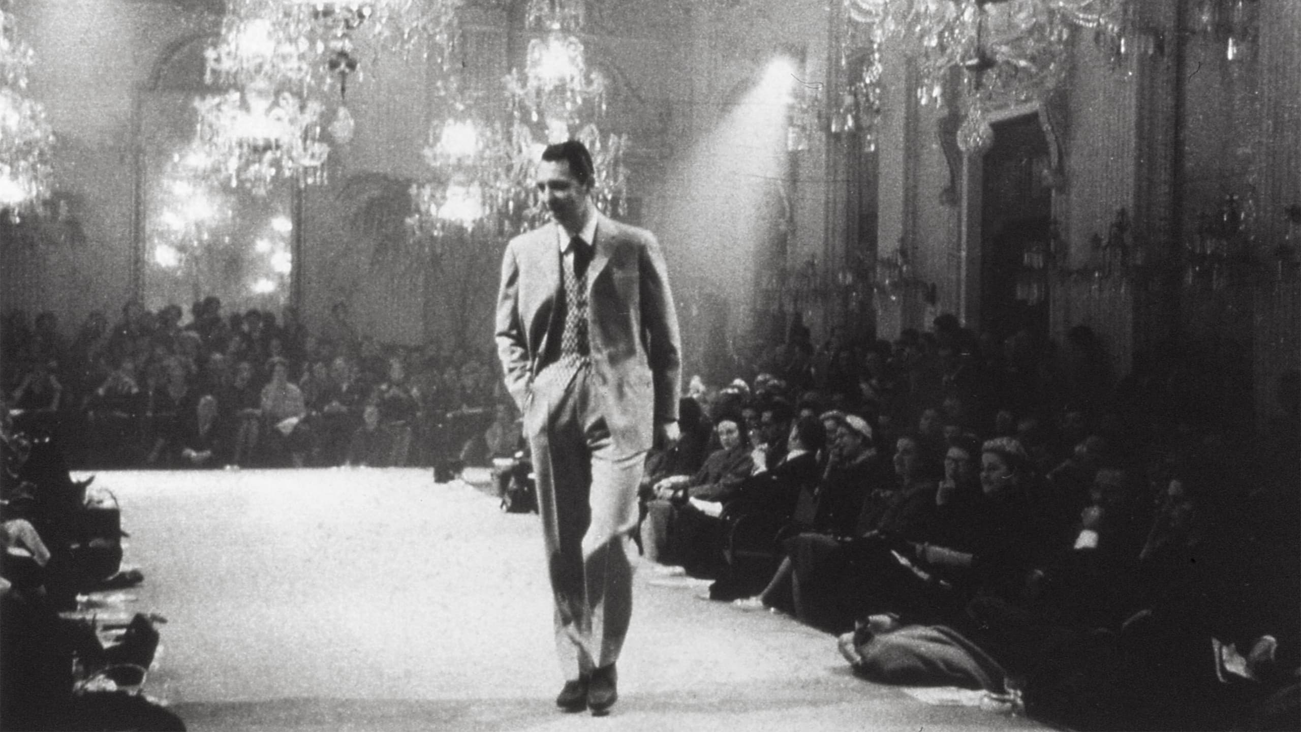 Man in Brioni suit walking on fashion runway with audience seated on both sides under elegant chandeliers in black and white photo.
