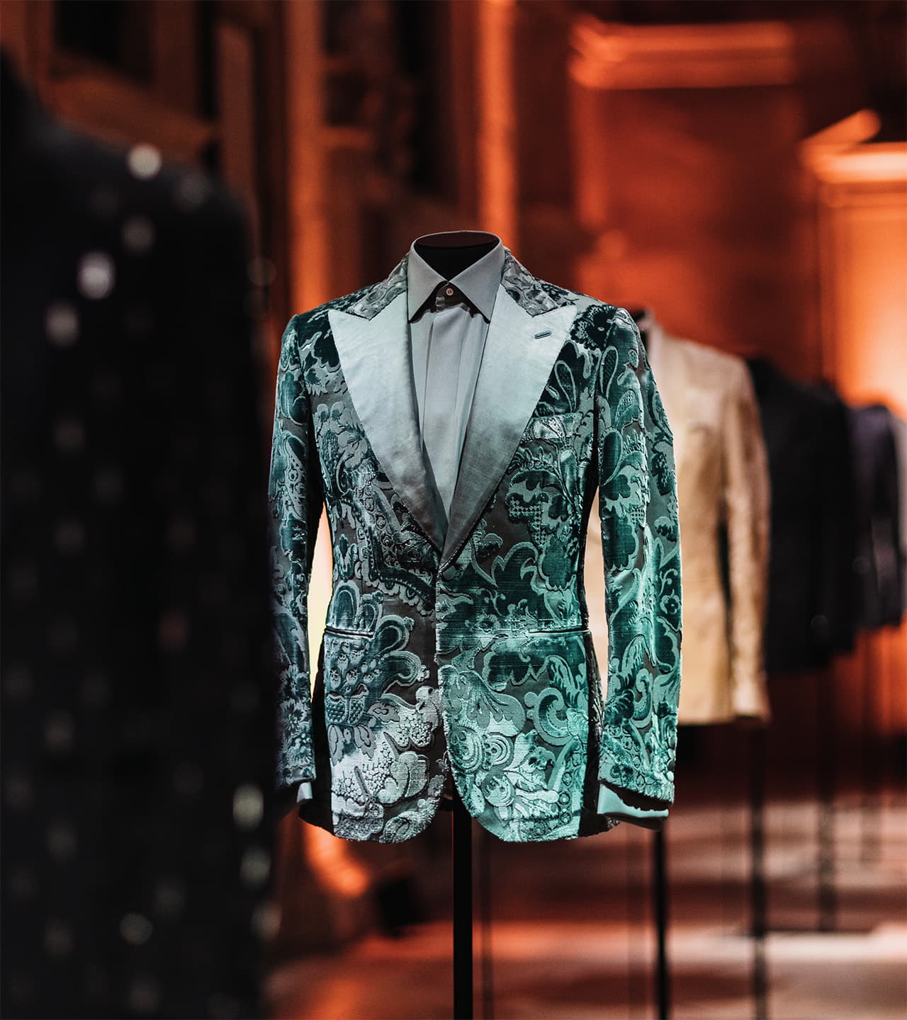 Brioni elegant teal velvet blazer with ornate floral pattern displayed on mannequin in luxury boutique with warm lighting.
