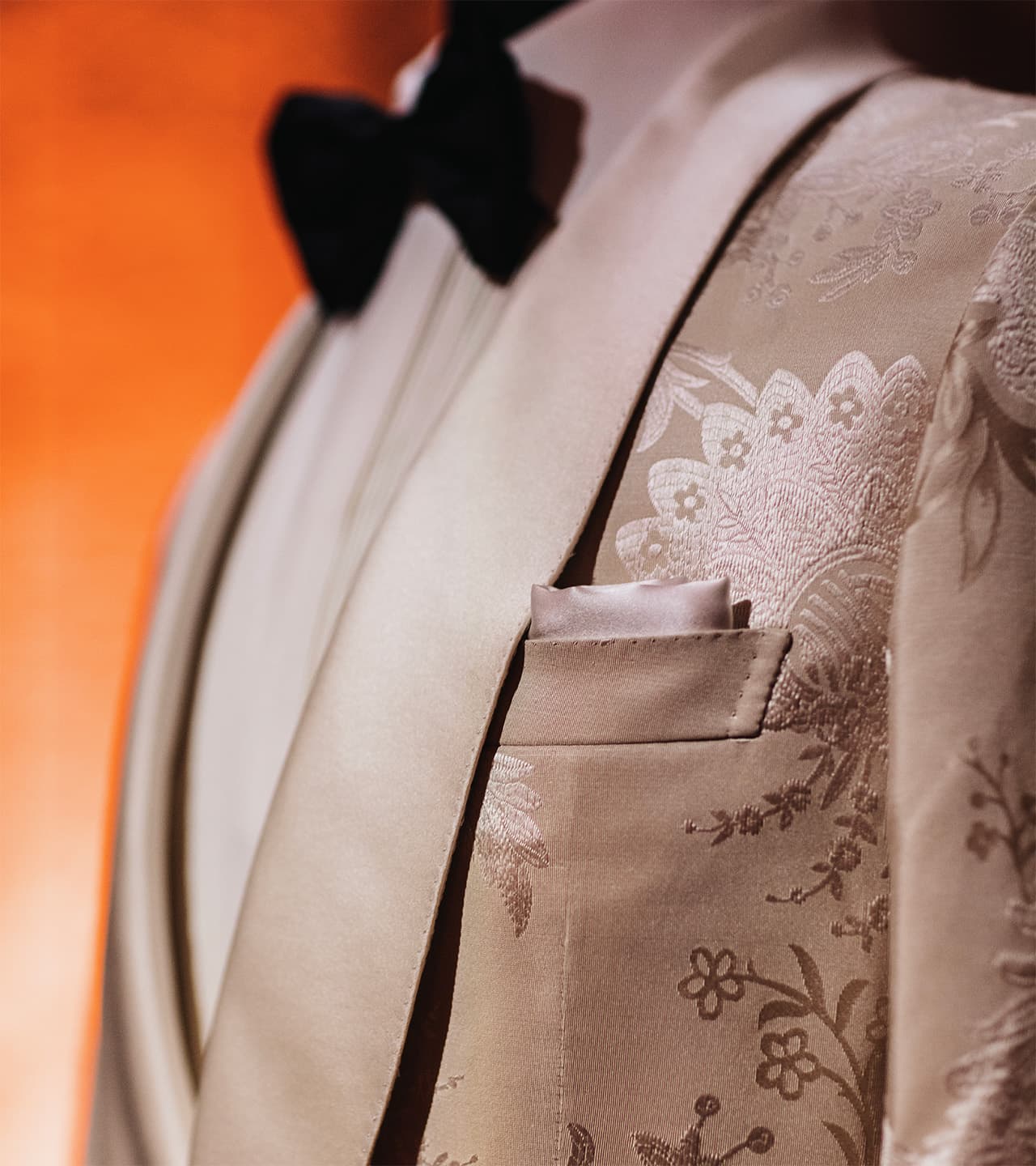 Close-up of Brioni's elegant cream tuxedo with floral pattern, black bow tie, and pocket square against orange background.