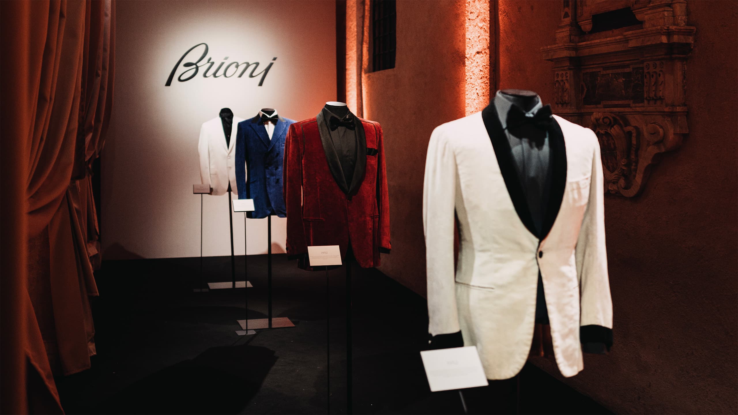 Brioni luxury tuxedo display featuring white, blue, and red velvet jackets with black lapels in an elegant showroom setting.