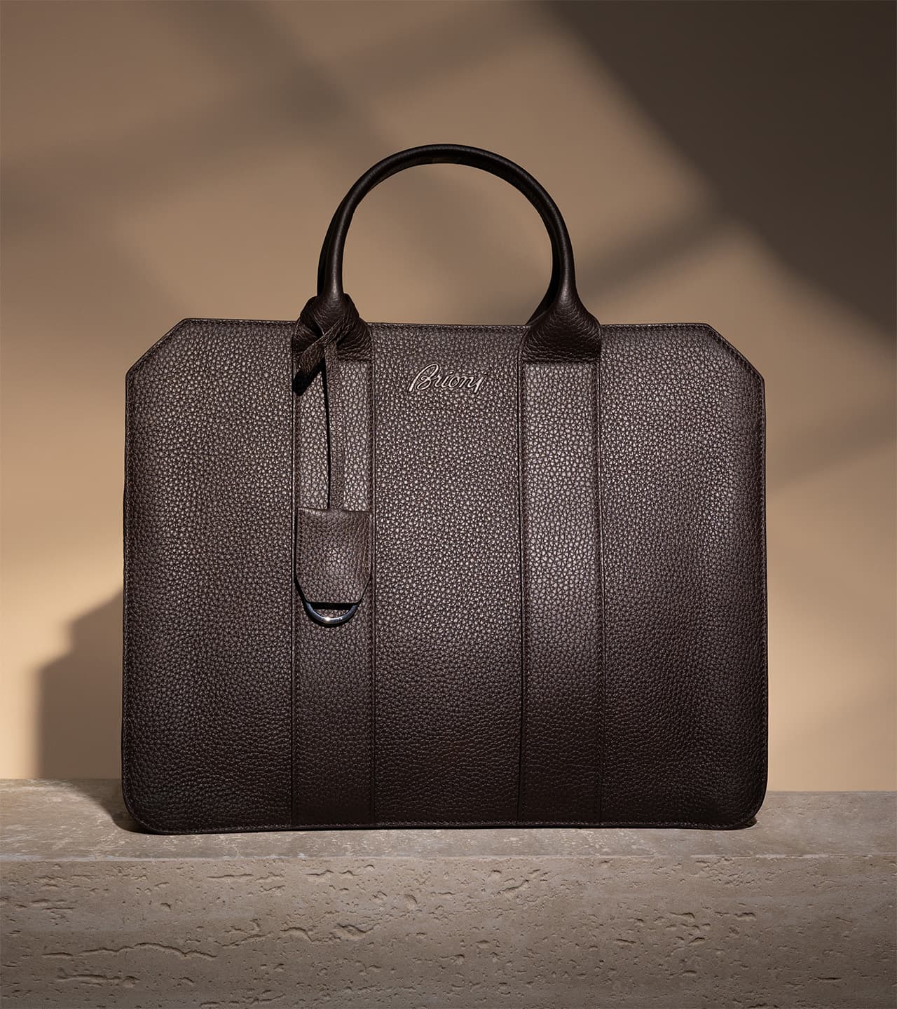 Brioni dark brown textured leather briefcase with dual handles and Brioni logo, displayed against a beige wall with shadows.