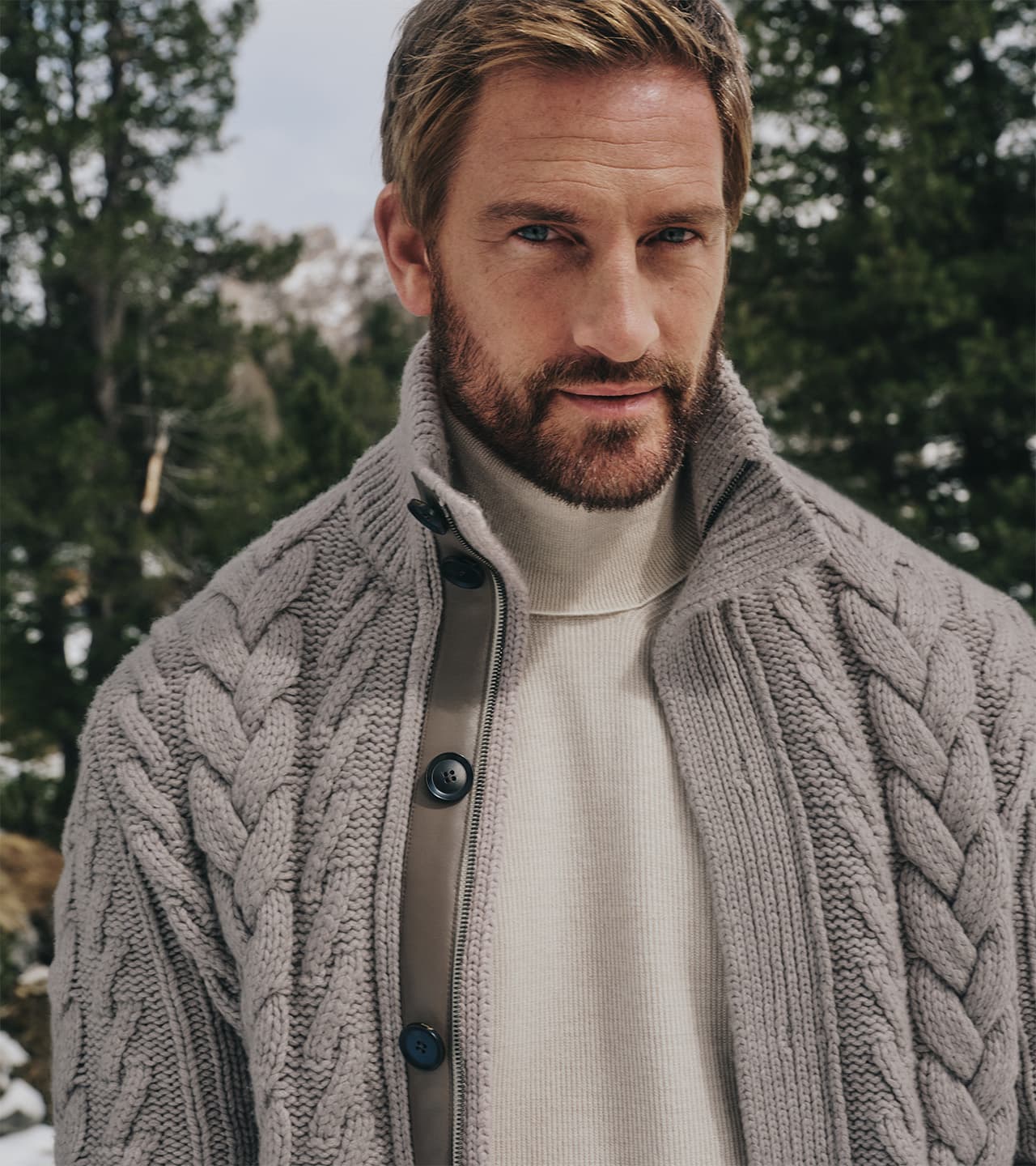 Person wearing a Brioni gray cable-knit cardigan over white turtleneck in a forest setting with evergreen trees behind.