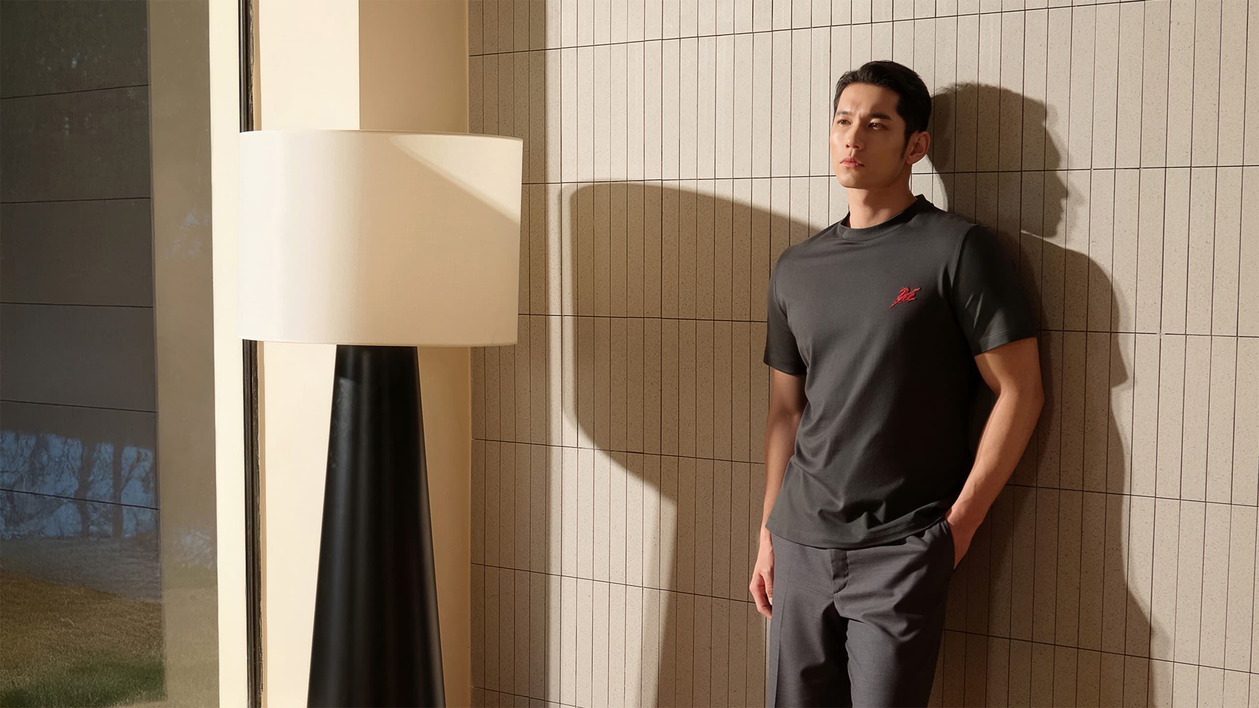 Man in Brioni dark gray t-shirt with red logo and gray pants leaning against white tiled wall near modern floor lamp.